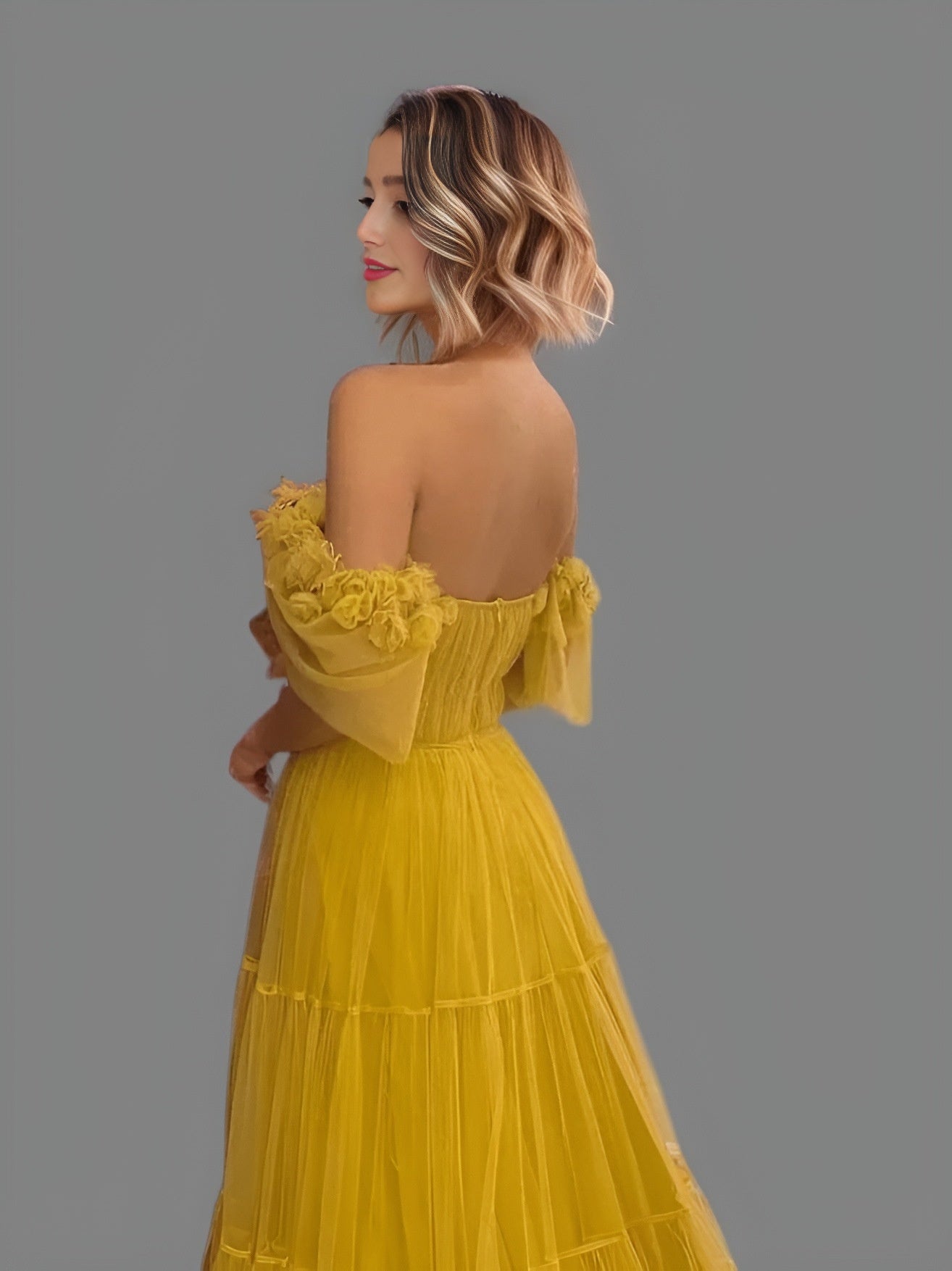 Woman wearing a formal gown with a 3D flowers around off the shoulder sleeves and an open back.