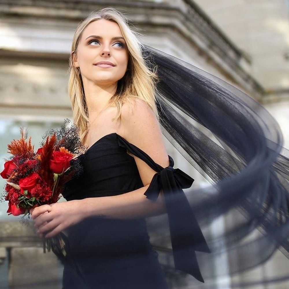 Bride in black dress standing outdoors with a red bouquet and a long black veil flowing around her in the wind.