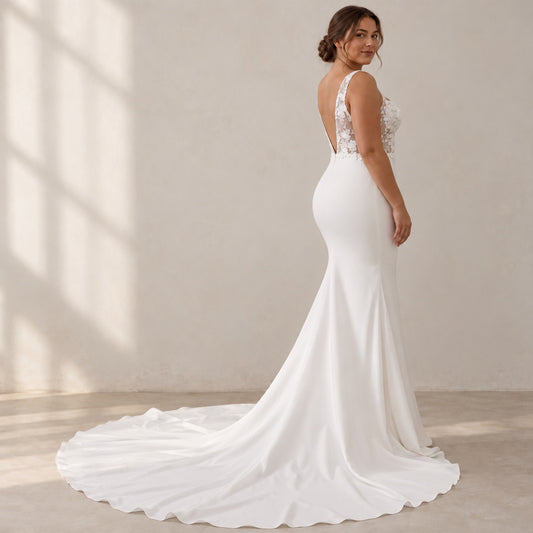 A woman modeling a white mermaid-style wedding gown with a fitted silhouette, lace-embellished back, and long flowing train in a softly lit indoor setting.