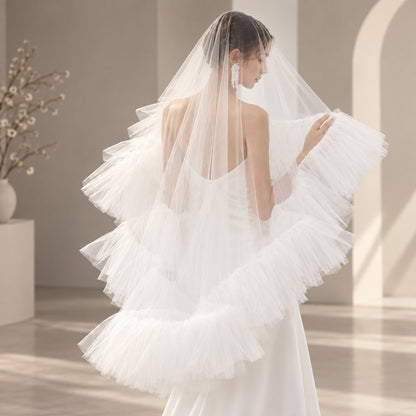 A woman wearing a long, layered tulle bridal veil and a white wedding dress, photographed from the back in a softly lit indoor setting with neutral walls and arches.
