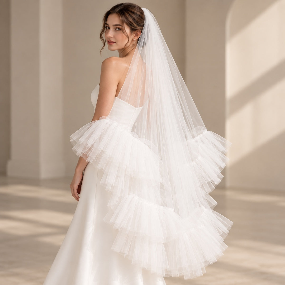 A woman wearing a bridal gown with a long layered tulle overskirt and matching long veil, photographed in a softly lit indoor setting with warm neutral walls and floor shadows.