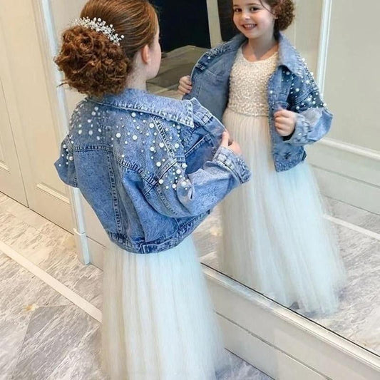 Young girl wearing a sparkly pearl denim jacket over a white dress, standing in front of a mirror.