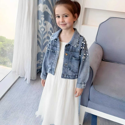 Young girl in a white dress and denim jacket standing in a grey room with a patterned curtain.