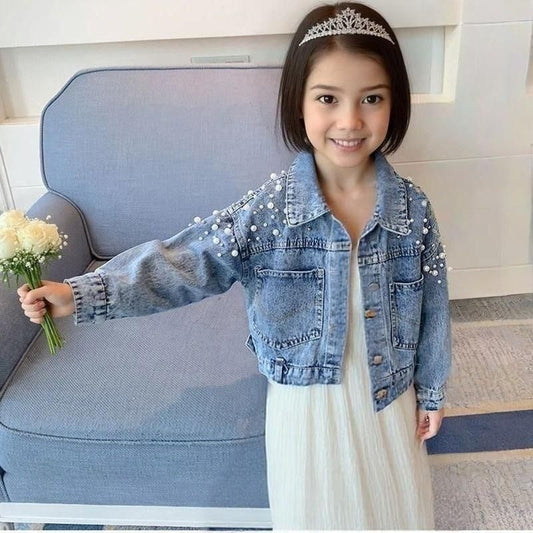 Young girl in a denim jacket with pearl embellishments and a tiara, holding flowers, sitting on a couch.