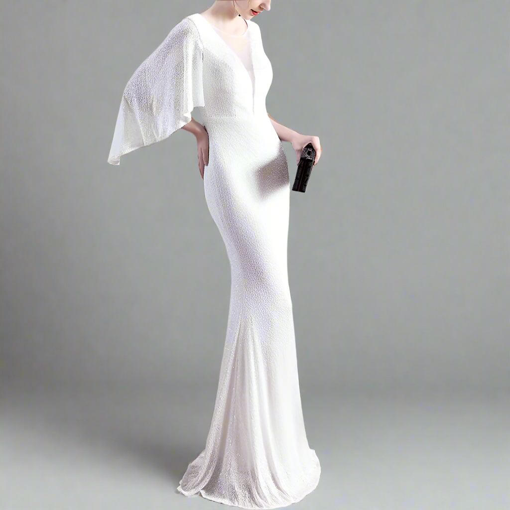 Woman in a white evening gown with a sheer overlay, holding a black clutch, standing against a neutral background.