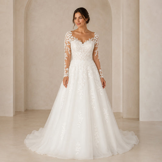 A woman modeling a long-sleeve white wedding gown with floral lace embroidery, standing in a softly lit beige interior with arched walls.
