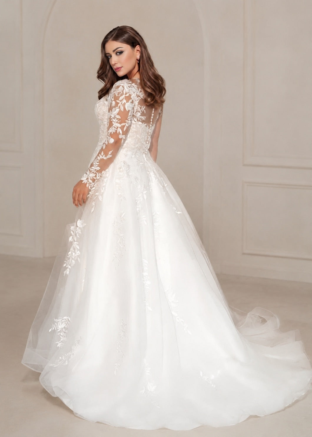 A woman modeling a long-sleeve white wedding gown with floral lace applique and a full tulle skirt, photographed in a softly lit cream-colored interior setting.