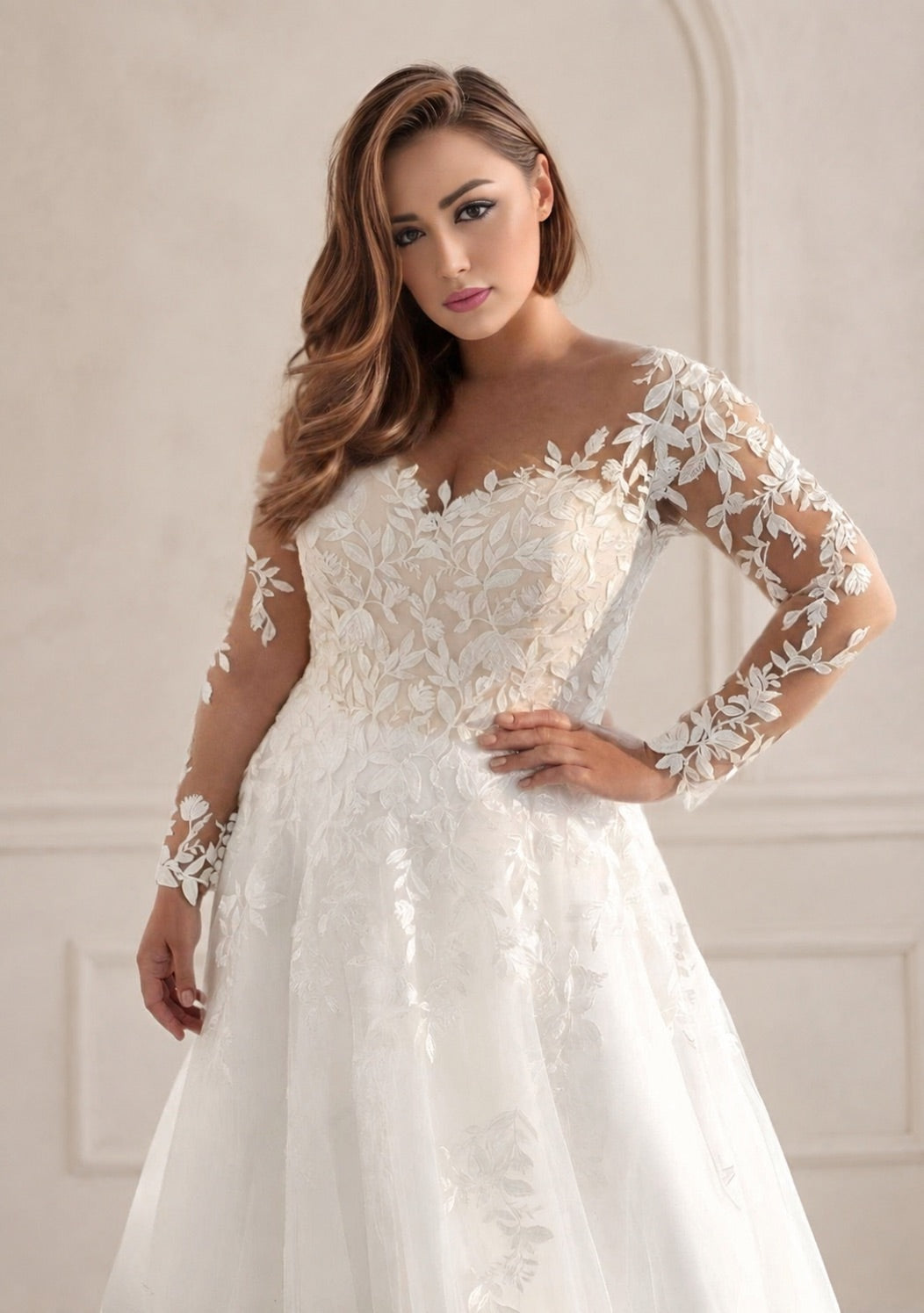 A woman modeling a white lace wedding gown with sheer long sleeves, a fitted lace bodice, and a full tulle skirt, posed against a soft beige interior wall.