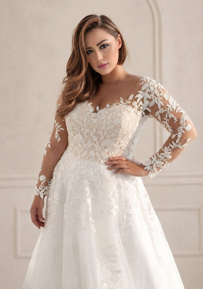 A woman modeling a white lace wedding gown with sheer long sleeves, a fitted lace bodice, and a full tulle skirt, posed against a soft beige interior wall.