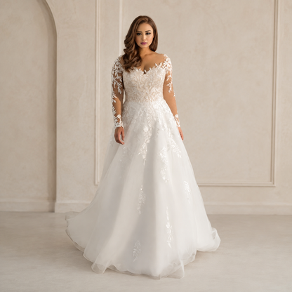 A woman modeling a white A-line wedding gown with long lace sleeves, a lace bodice, and a full tulle skirt, standing in a softly lit cream-colored interior with arched wall details.