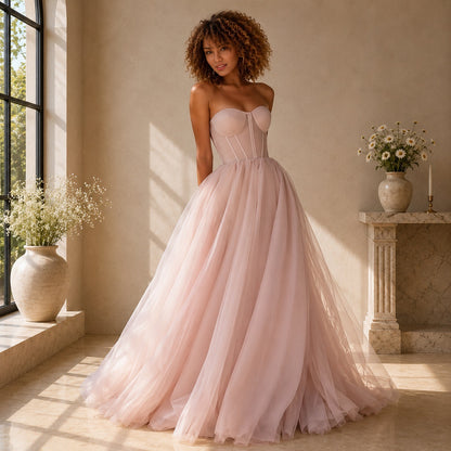 A woman stands indoors wearing a strapless blush pink ball gown with a fitted corset-style bodice and a full tulle skirt. The room has warm beige walls, a window on the left, and a stone mantel with flowers on the right.