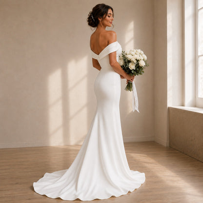 A woman wearing an off-the-shoulder white wedding gown with a fitted silhouette and a long train, standing in a sunlit room and holding a bouquet of white flowers.