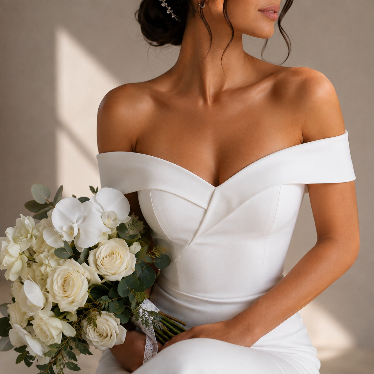 A woman wearing an off-the-shoulder white wedding dress with structured seams, holding a bouquet of white roses and greenery against a softly lit neutral background.
