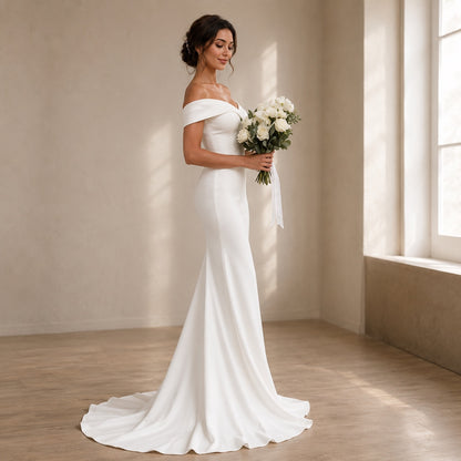 A woman wearing an off-the-shoulder white wedding gown with a fitted silhouette and long train, standing in a sunlit room and holding a bouquet of white flowers.