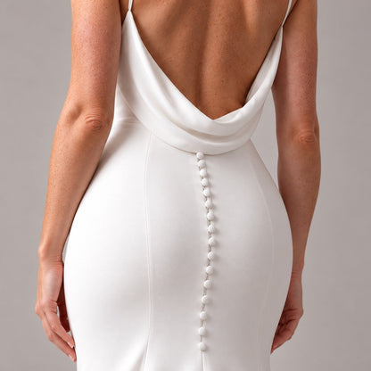 Close-up back view of a woman wearing a white fitted dress with a draped open back and a vertical row of decorative buttons down the center, against a plain light gray studio background.