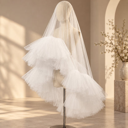 A long white bridal veil with layered tulle ruffles displayed on a mannequin head in a softly lit indoor setting with warm beige walls and shadows.