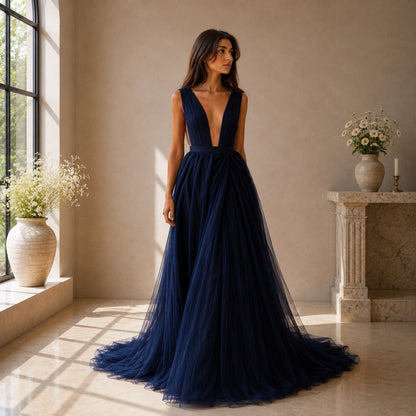 A woman stands in a sunlit interior wearing a floor-length navy gown with a deep V-neckline, fitted bodice, and voluminous tulle skirt. The room has beige walls, a stone fireplace, and large windows with flower arrangements.
