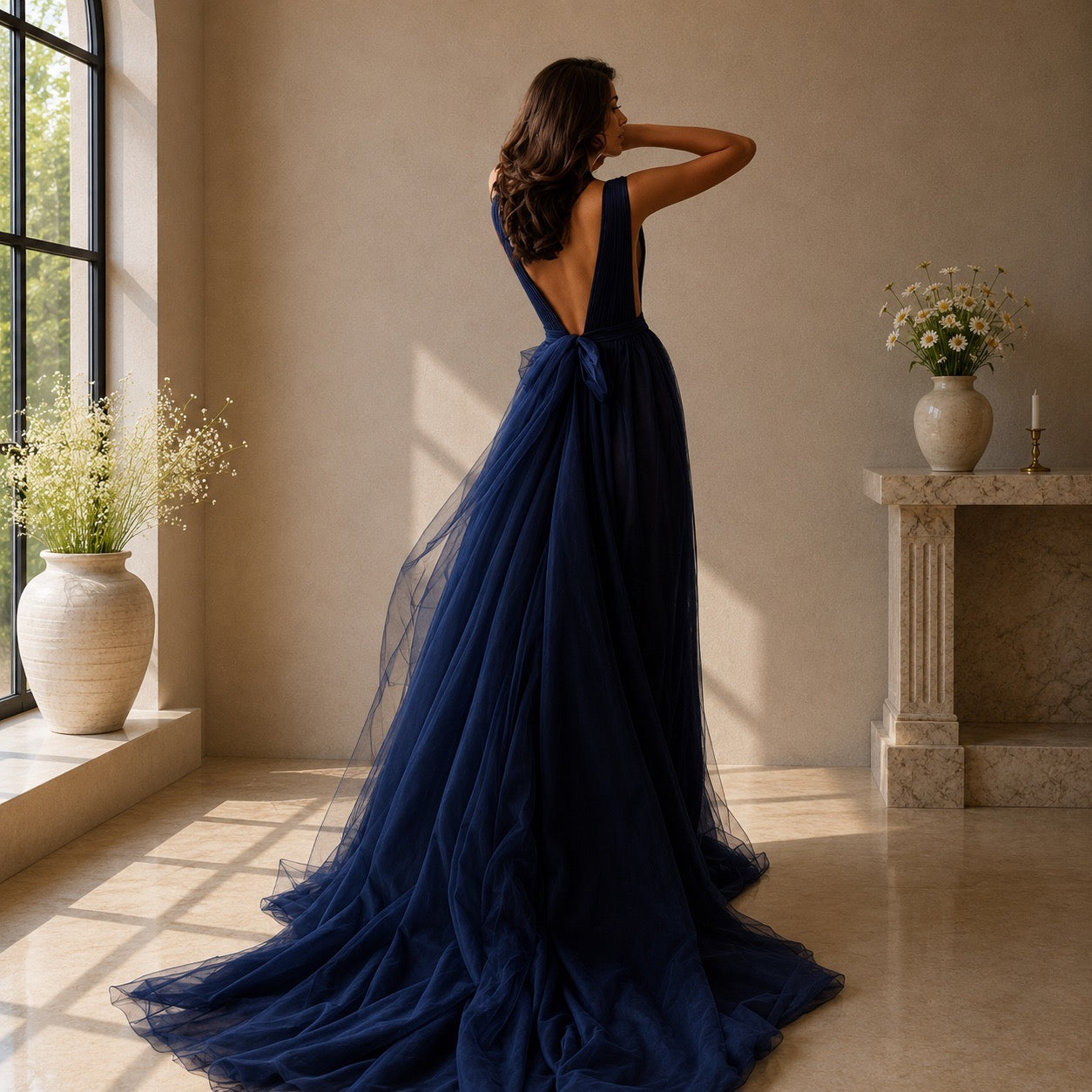 A woman stands with her back to the camera wearing a floor-length navy blue tulle gown with a deep open back and a large bow at the waist, photographed in a sunlit indoor room with a window, vases, and a stone mantel.