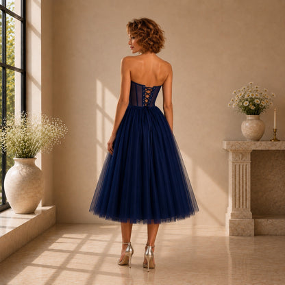 A woman stands with her back to the camera wearing a strapless navy blue tulle dress with a lace-up corset-style bodice, in a sunlit indoor room with beige walls and decorative vases.