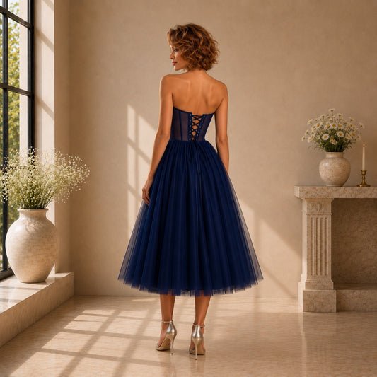 A woman stands with her back to the camera wearing a strapless navy blue tulle dress with a lace-up corset-style bodice, in a sunlit indoor room with beige walls and decorative vases.