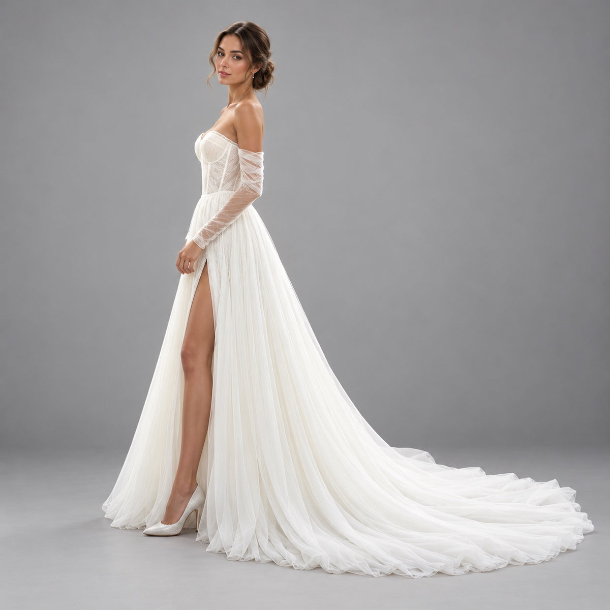 A woman modeling an off-the-shoulder white wedding gown with sheer long sleeves, a high leg slit, and a flowing train against a plain gray studio background.