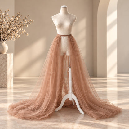 A long, flowing tulle skirt in a dusty blush pink color displayed on a cream mannequin dress form, set in a warm, sunlit room with beige walls and a marble floor.