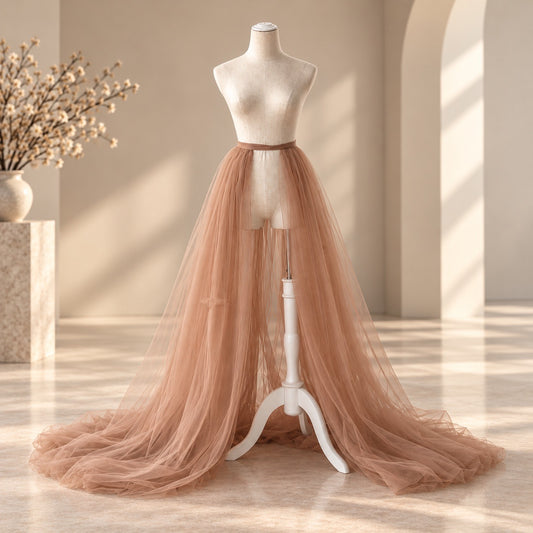 A long, flowing tulle skirt in a dusty blush pink color displayed on a cream mannequin dress form, set in a warm, sunlit room with beige walls and a marble floor.