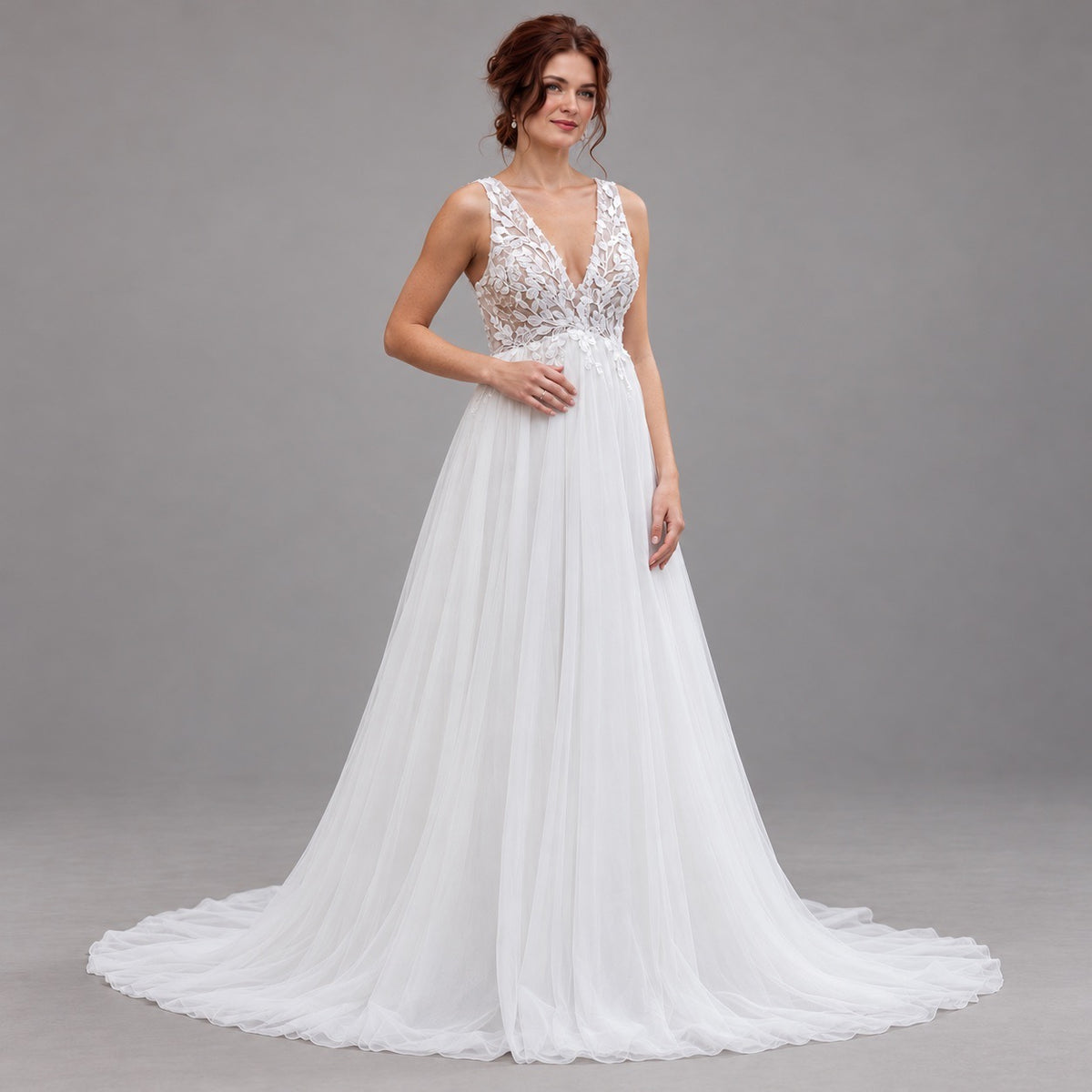 A woman modeling a white wedding gown with a lace bodice, deep V-neckline, and a flowing tulle skirt with a train, photographed against a plain gray studio background.