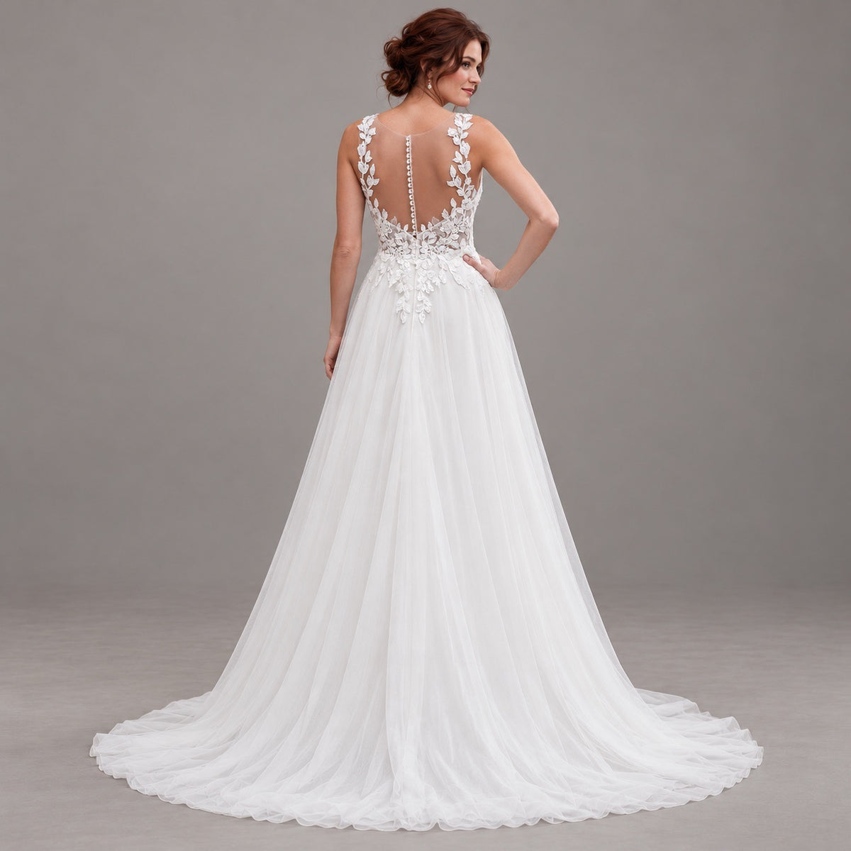 A woman modeling a white wedding gown from the back, showing a deep V-back with floral lace applique, a beaded waistband, and a full tulle train against a neutral gray studio background.