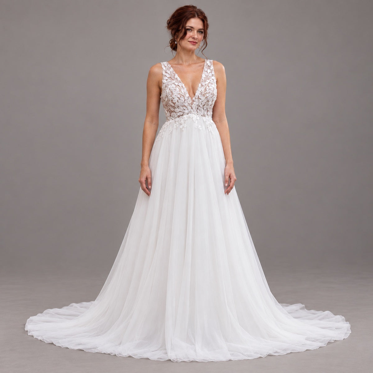 A woman modeling a white wedding gown with a lace bodice, deep V-neckline, and a full flowing tulle skirt with a train, photographed against a plain gray studio background.