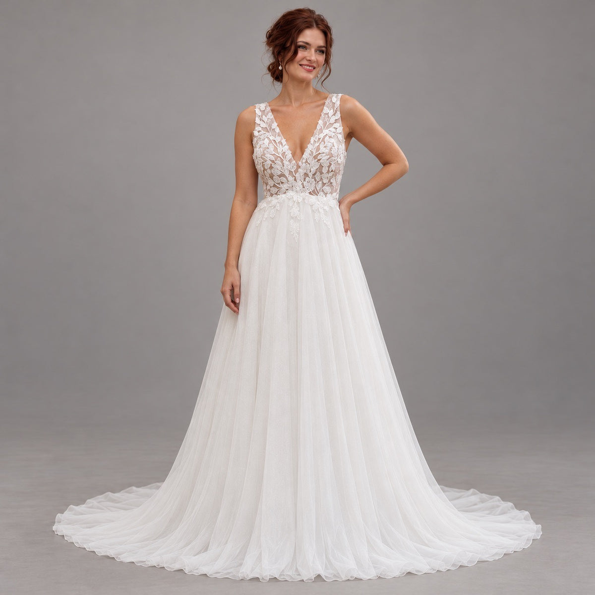 A woman modeling a white wedding gown with a lace V-neck bodice, sheer straps, and a full tulle skirt with a train, photographed against a plain gray studio background.