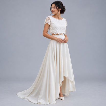 A woman models a two-piece ivory wedding dress with a lace crop top and a flowing high-low satin skirt against a neutral gray studio background.