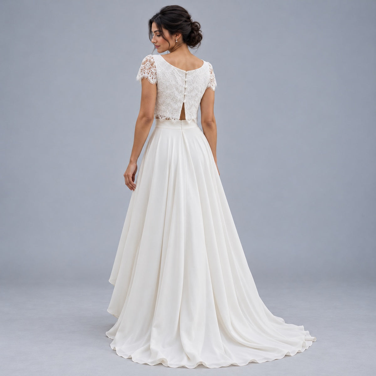 A woman modeling a white wedding dress from the back, featuring a lace cap sleeves, open back, and a flowing chiffon A-line skirt with a train, set against a solid gray studio background.