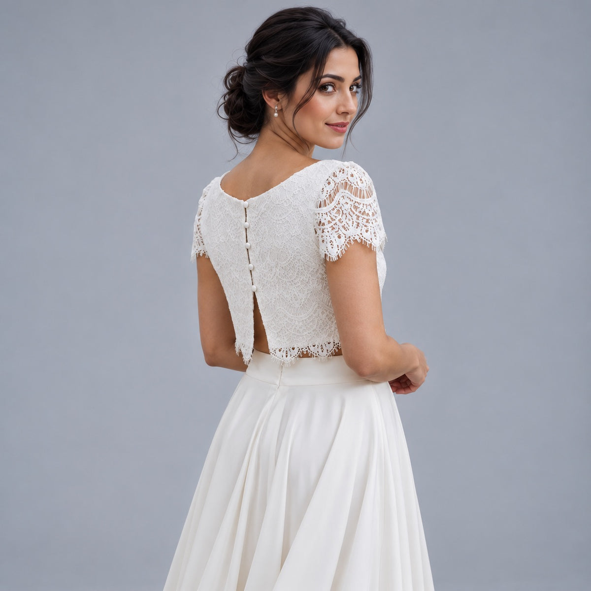 A woman wearing a white lace and satin bridal gown, shown from the back as she looks over her shoulder against a solid gray studio background.