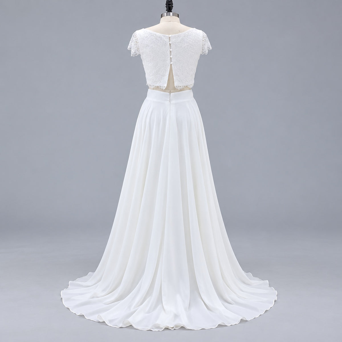 A white two-piece formal gown displayed on a mannequin against a light gray studio background. The dress features a lace short-sleeve crop top with a back zipper and a flowing chiffon skirt with a slight train.