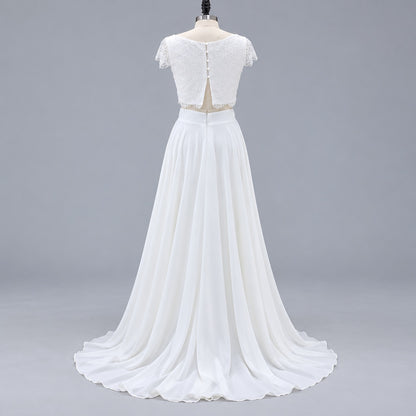 A white two-piece formal gown displayed on a mannequin against a light gray studio background. The dress features a lace short-sleeve crop top with a back zipper and a flowing chiffon skirt with a slight train.