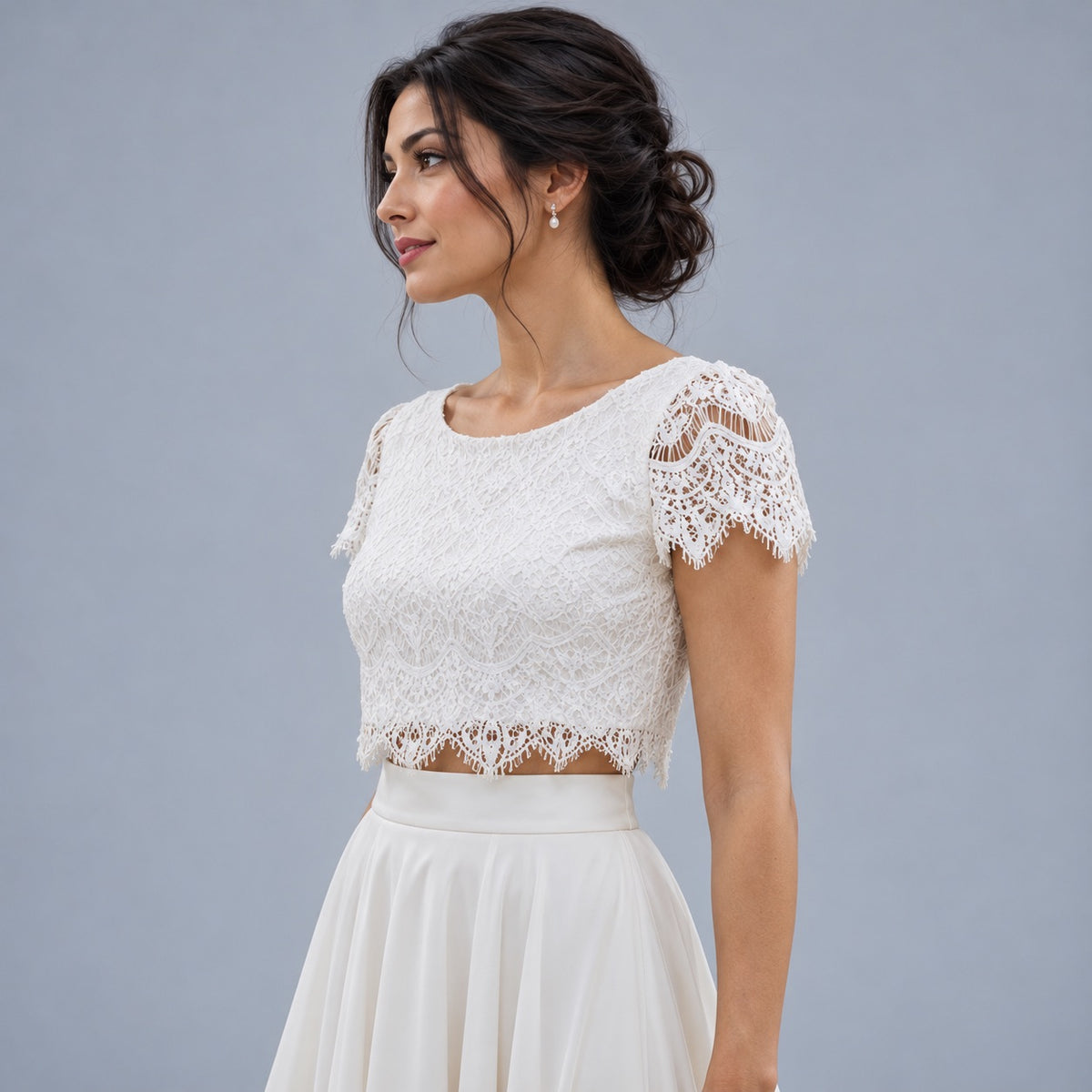 A woman wearing a white lace short-sleeve crop top with a flared skirt, posed against a solid gray studio background.