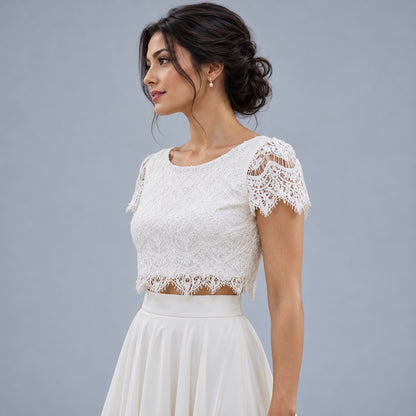 A woman wearing a white lace short-sleeve crop top with a flared skirt, posed against a solid gray studio background.