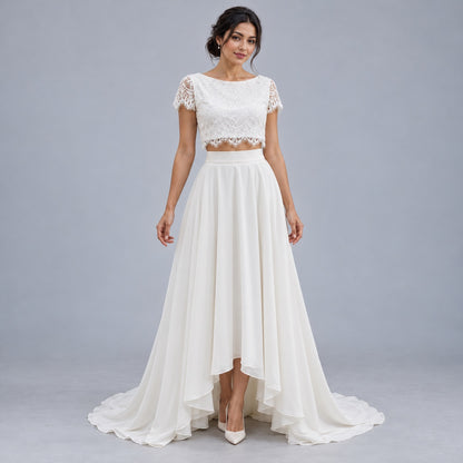A woman modeling a two-piece white bridal outfit consisting of a short-sleeve lace crop top and a high-low flowing skirt, photographed against a neutral gray studio background.
