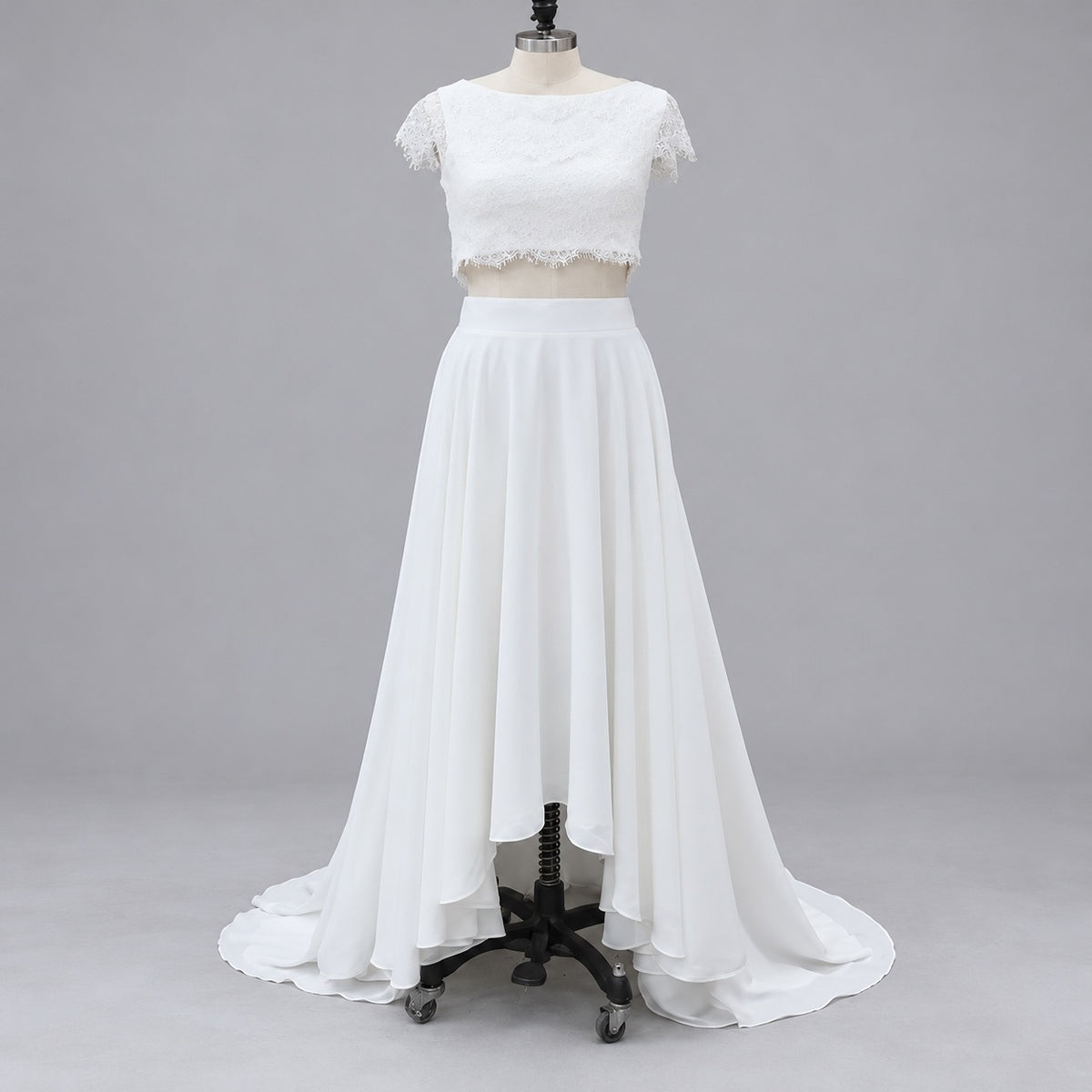 White two-piece formal gown displayed on a dress form mannequin against a neutral gray studio background. The outfit features a short-sleeve lace crop top with scalloped edges and a high-waisted, flowing chiffon maxi skirt with a slight train.