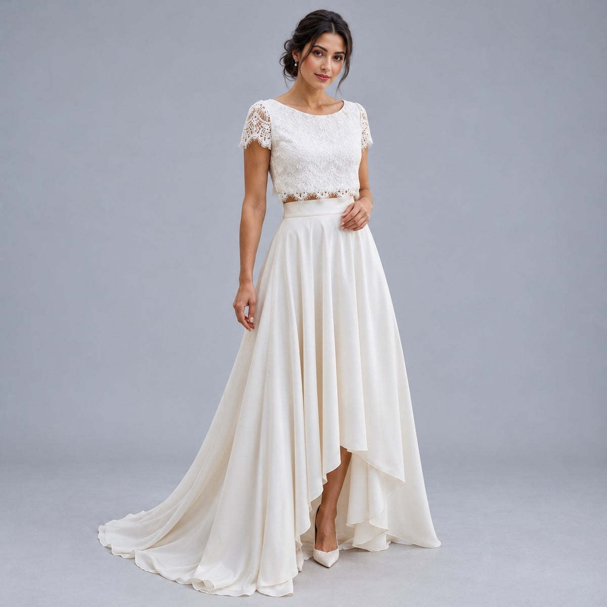 A woman modeling a long cream bridal gown with a lace embroidered bodice, short lace sleeves, and a flowing high-low skirt against a solid gray studio background.