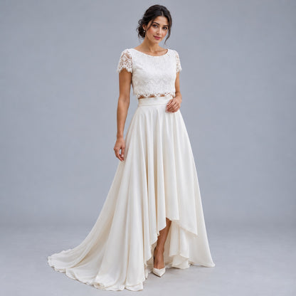 A woman modeling a long cream bridal gown with a lace embroidered bodice, short lace sleeves, and a flowing high-low skirt against a solid gray studio background.
