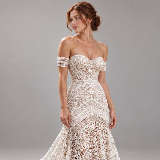 An off-the-shoulder lace wedding gown in an ivory/cream tone, featuring intricate floral embroidery and a sweetheart neckline, modeled against a solid gray studio background.