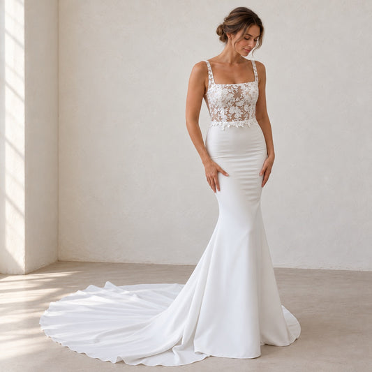 A woman modeling a white mermaid-style wedding gown with a lace bodice, square neckline, thin straps, and a long train in a softly lit indoor room with beige walls and floor.