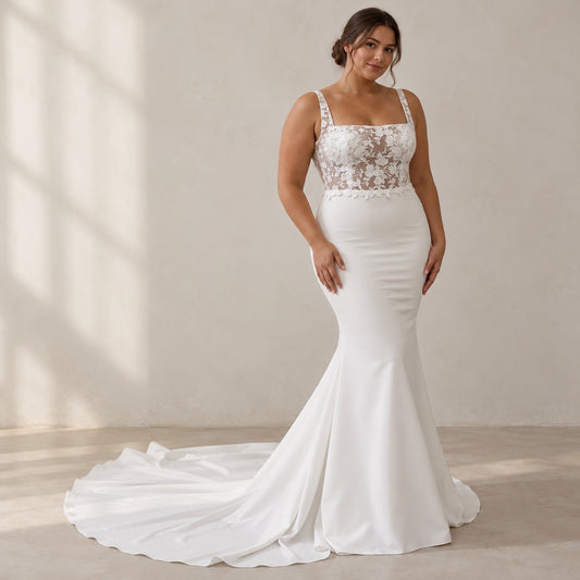 A woman modeling a white mermaid-style wedding dress with a lace bodice and thin straps, standing in a softly lit indoor studio setting with a long train on the floor.
