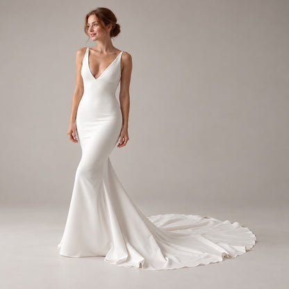 A woman modeling a white sleeveless mermaid-style wedding gown with a deep V-neckline and a long flowing train against a plain light gray studio background.
