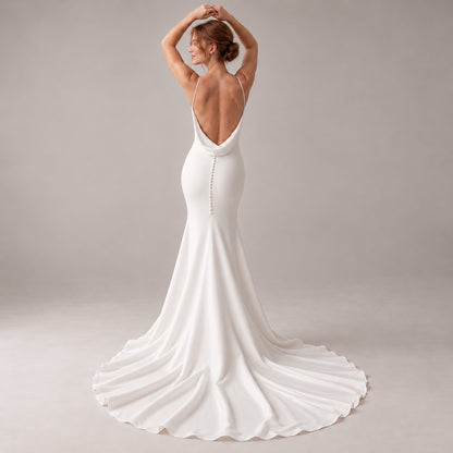 A woman wearing a white bridal gown is photographed from the back against a soft light gray studio background. The dress has thin straps, a deep open back, and a long train with a slight train wave.