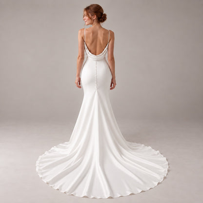 A woman modeling a white bridal gown from the back, showing a deep V-back, thin straps, and a long satin train against a neutral studio backdrop.
