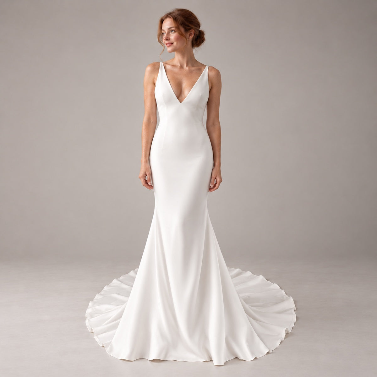 A woman modeling a sleeveless white mermaid-style wedding gown with a deep V-neckline and a full skirt train, photographed against a plain light gray studio background.