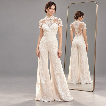 A woman models a cream lace high-neck jumpsuit with short sleeves and wide flared legs, standing in front of a floor-to-ceiling mirror that shows the back view of the dress. The background is a clean, neutral gray studio setting.